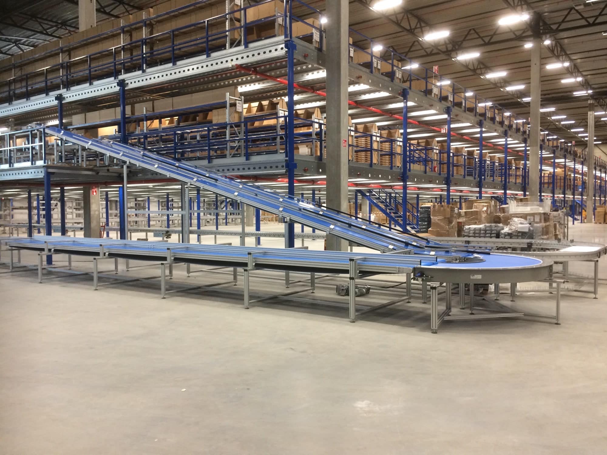 Transportbanden of riemtransport die producten van op verschillende hoogtes vervoert. De lijn staat in een grote productiehal of een magazijn en heeft een blauwe band. Dynamic Conveyor Solutions zorgt voor de engineering van conveyor systemen om soortgelijke oplossingen te creëren en staat ook in voor het onderhoud van conveyors.